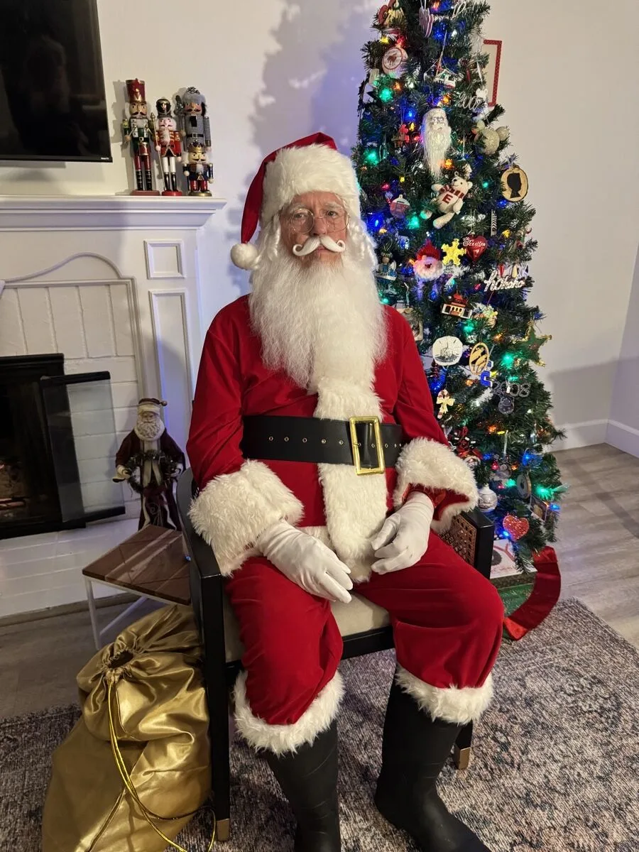 Santa seated beside a lit holiday tree and fireplace, full suit and boots visible