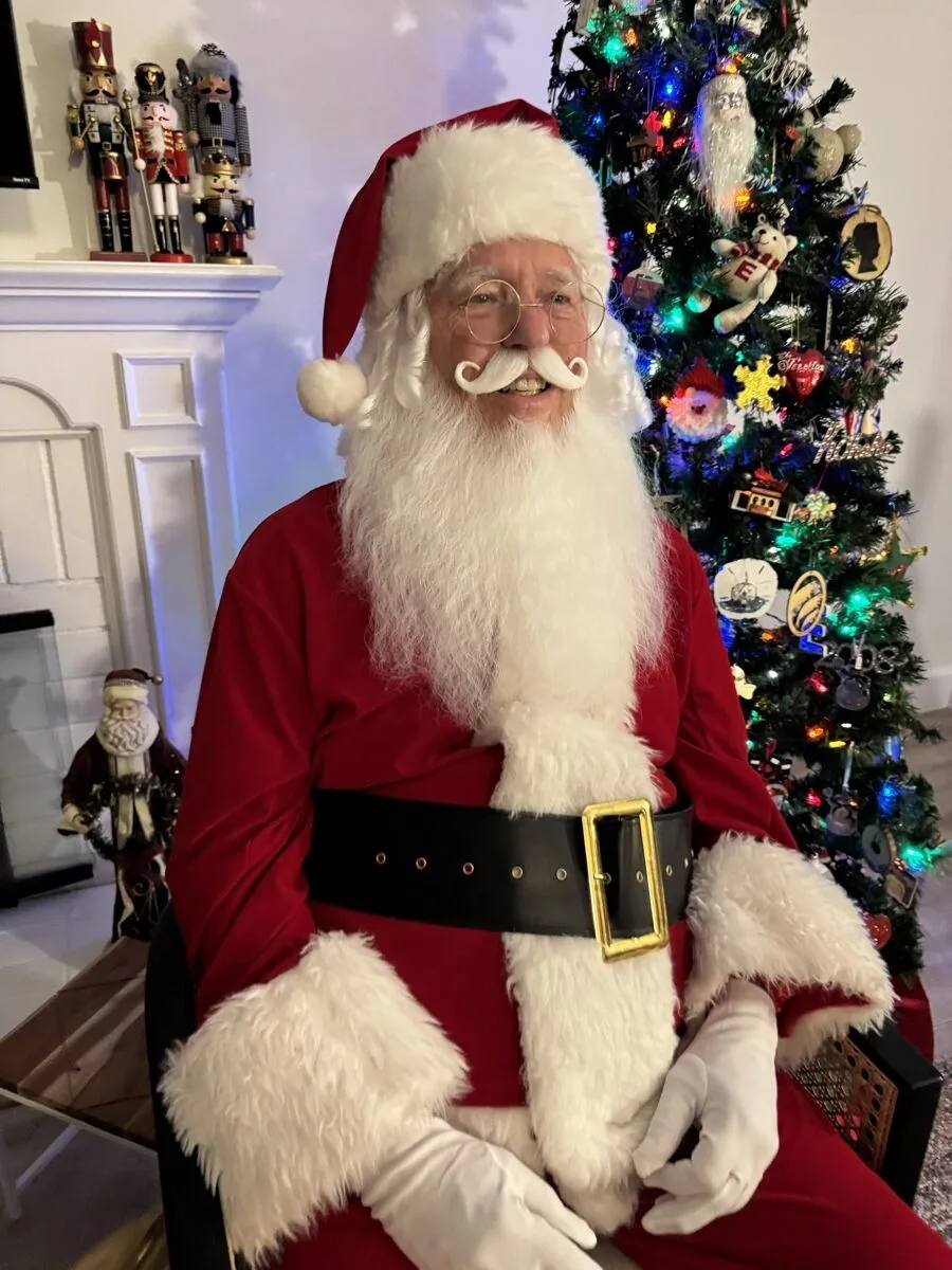 Santa in a red velvet suit and white beard seated in front of a decorated holiday tree