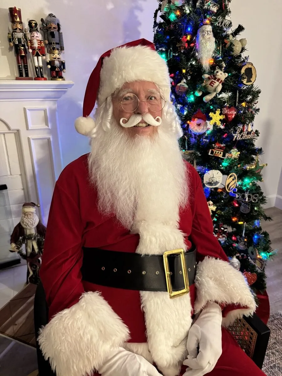 Santa smiling in his red velvet suit with white fur trim beside a decorated tree