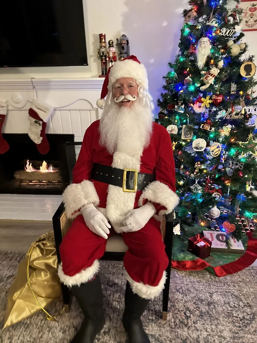 Santa seated by a fireplace with stockings and a decorated tree behind him
