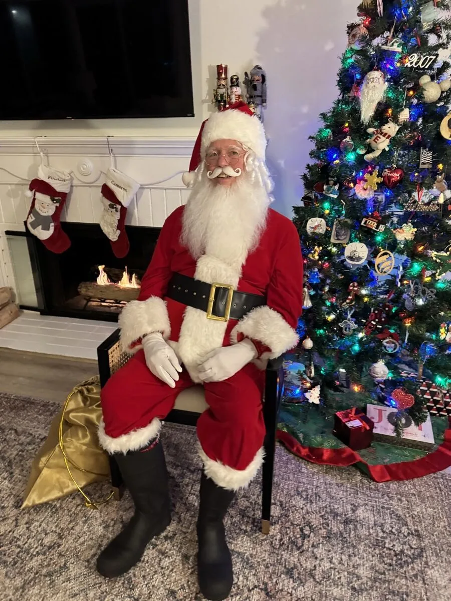 Santa seated in front of a fireplace and a lit holiday tree, with a gold sack of gifts beside him