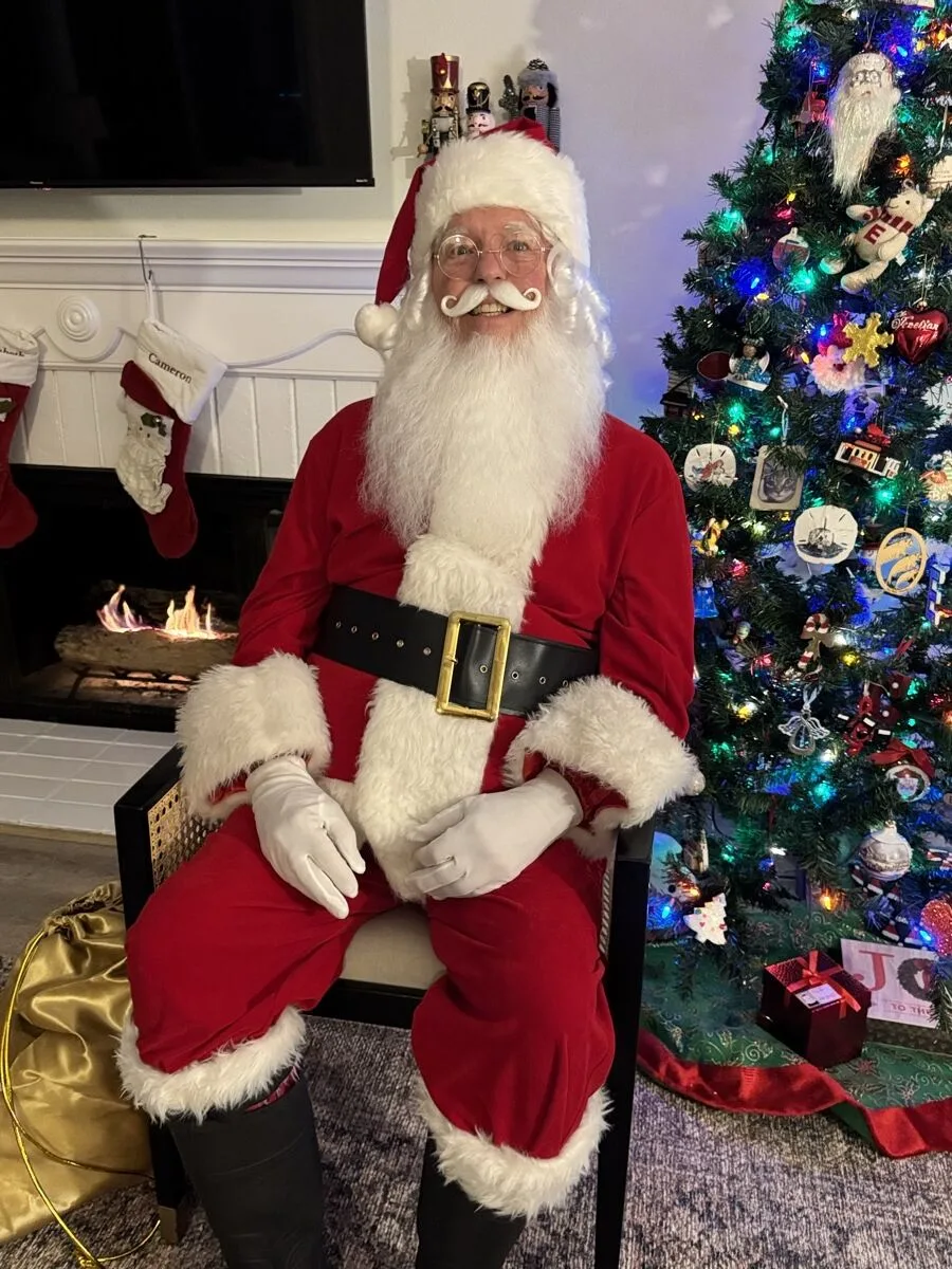 Santa with gloved hands resting on his knees in front of a glowing tree